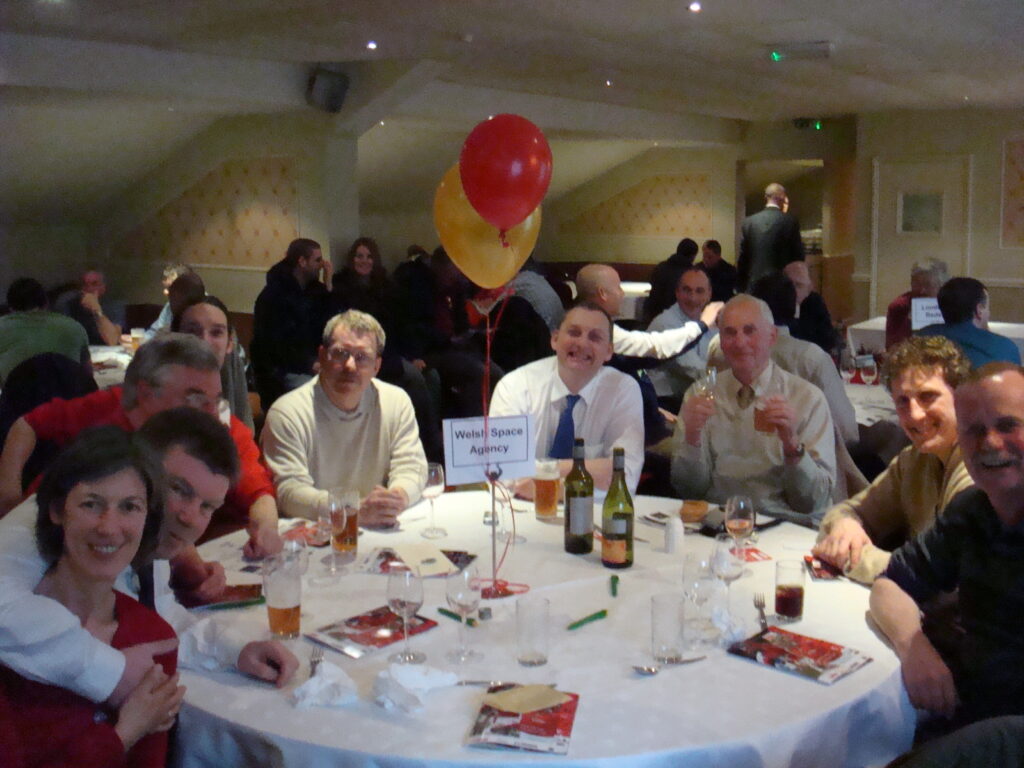 A group photo of Welsh Space Agency staff in a busy club house around a table with a white cloth and drinks on the table. With two balloons floating from the middle.