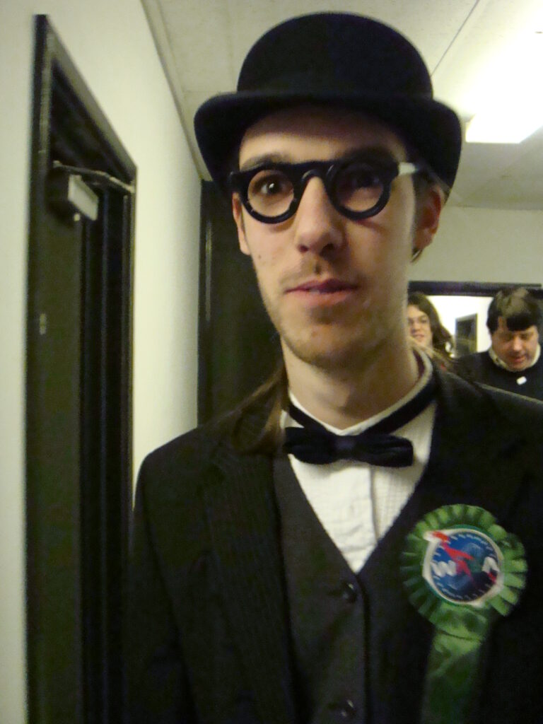 A man with a black suit, dickie bow tie, bowler hat and spectacles wearing a green rosette with the WASA logo on it.