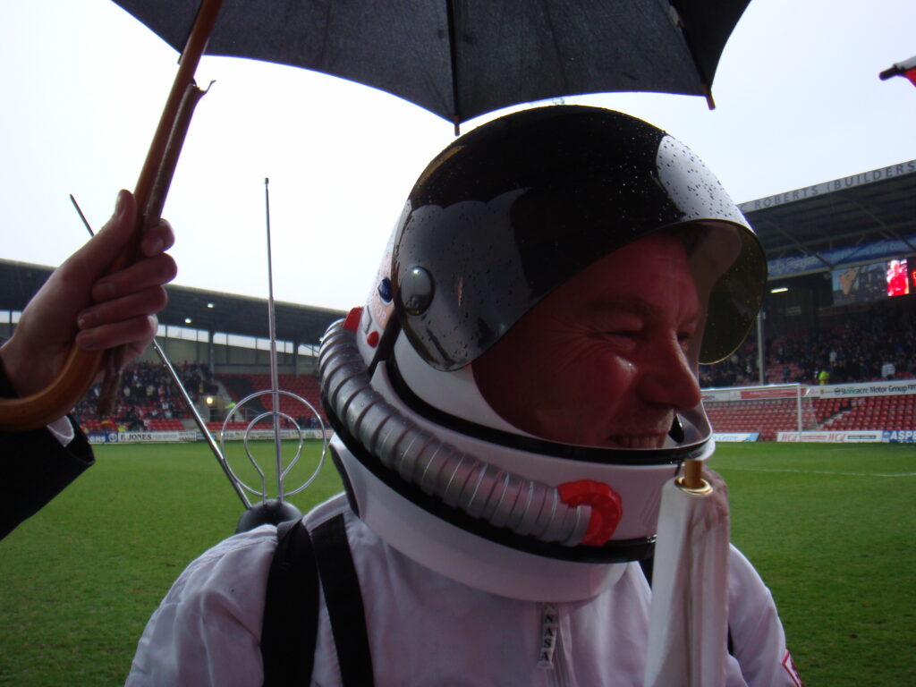 A smiling man in a spacesuit and helmet on the pitch with his assistant holding a brolley over him.