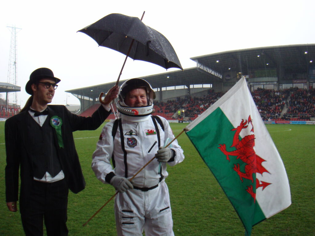 Man in spacesuit in the rain on the edge of the pitch before the match. He holds a welsh flag and his bowler hatted, bow-tied assistant hold an umbrella over his head.