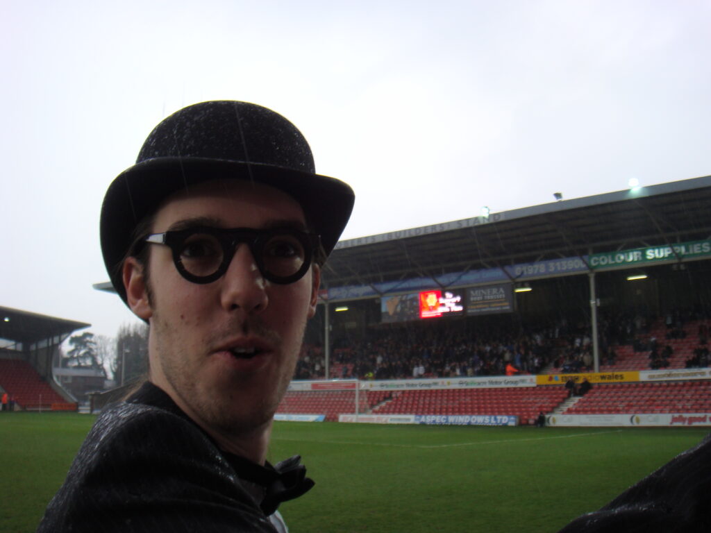 A tall man in bowler hat and spectacles in front of a football stand. he looks surprised