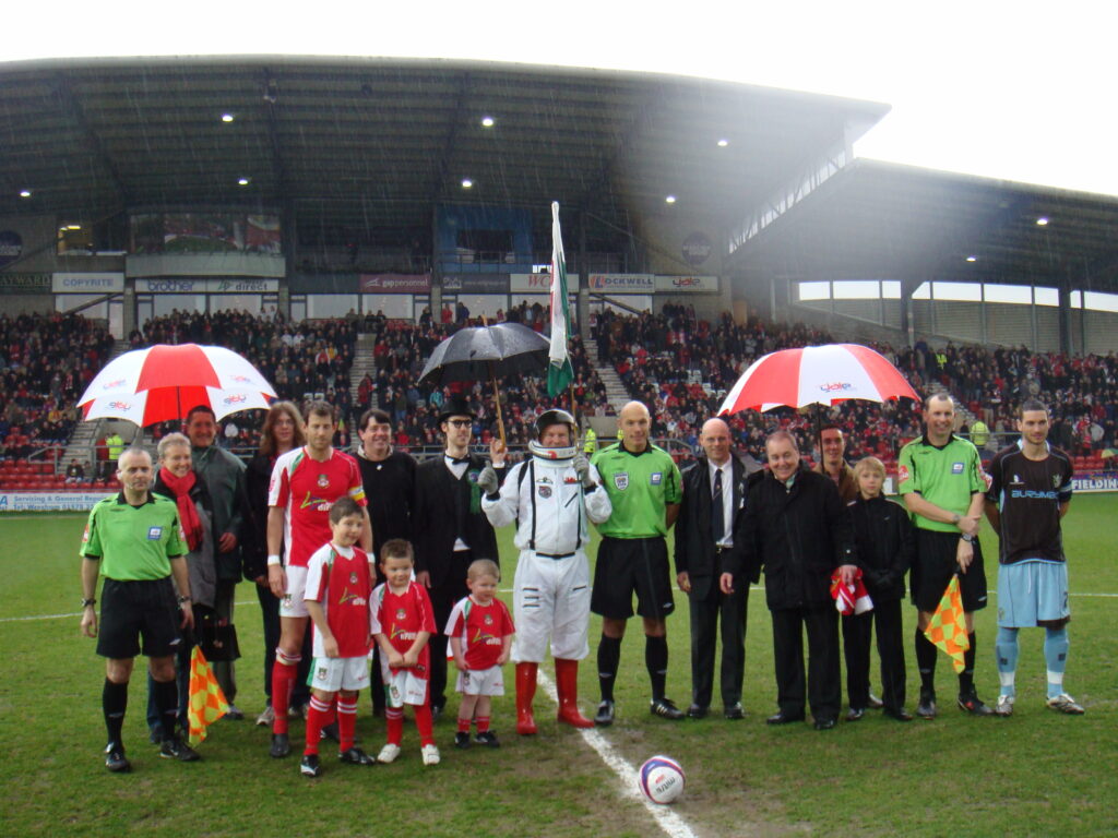 Match sponsors and officials line up for photo shoot in pitch centre on rainy day. Centre of photo is man in an astronaut suit next to referee Howard Webb.