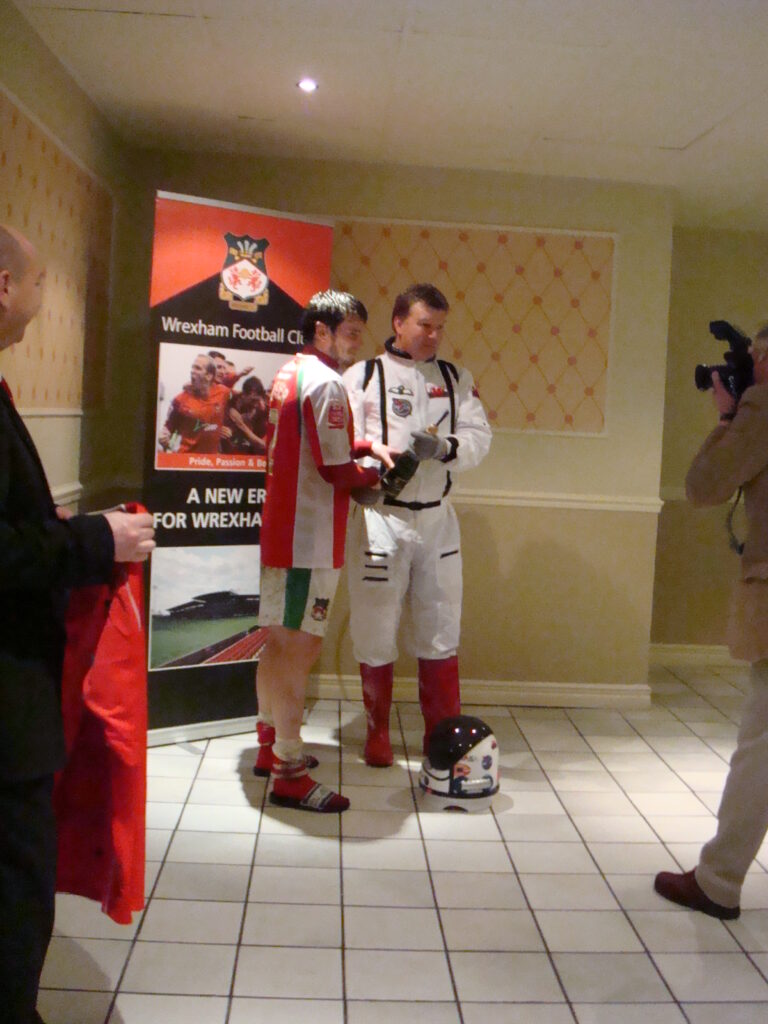 A man in a spacesuit hands a bottle of fizz to a footballer in the corner of a club house.
