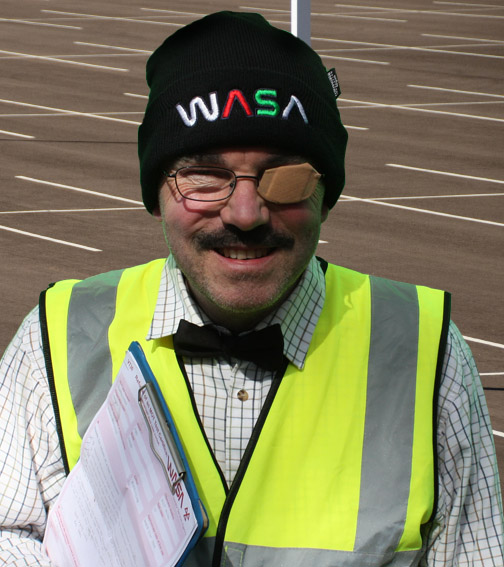 Morris Spoon wearing a WASA hat and a hi-viz vest, holding a clipboard in an empty car park.