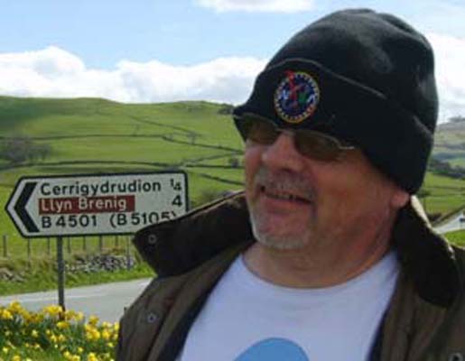 Plump man with sunglasses and a WASA hat stood in front of a Cerrigydrudion road sign.