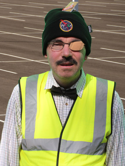 Man with hi-viz coat, shirt and tie and black WASA hat. Wearing spectacles with sticking plaster over one eye.