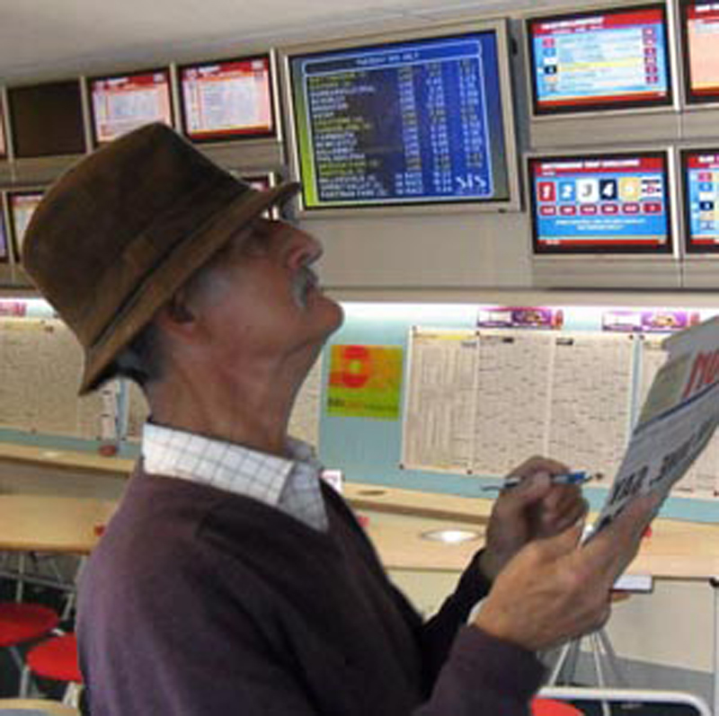 A middle aged man in a trilby at a betting shop studies the form on the many tv screens.  He is holding a newspaper.